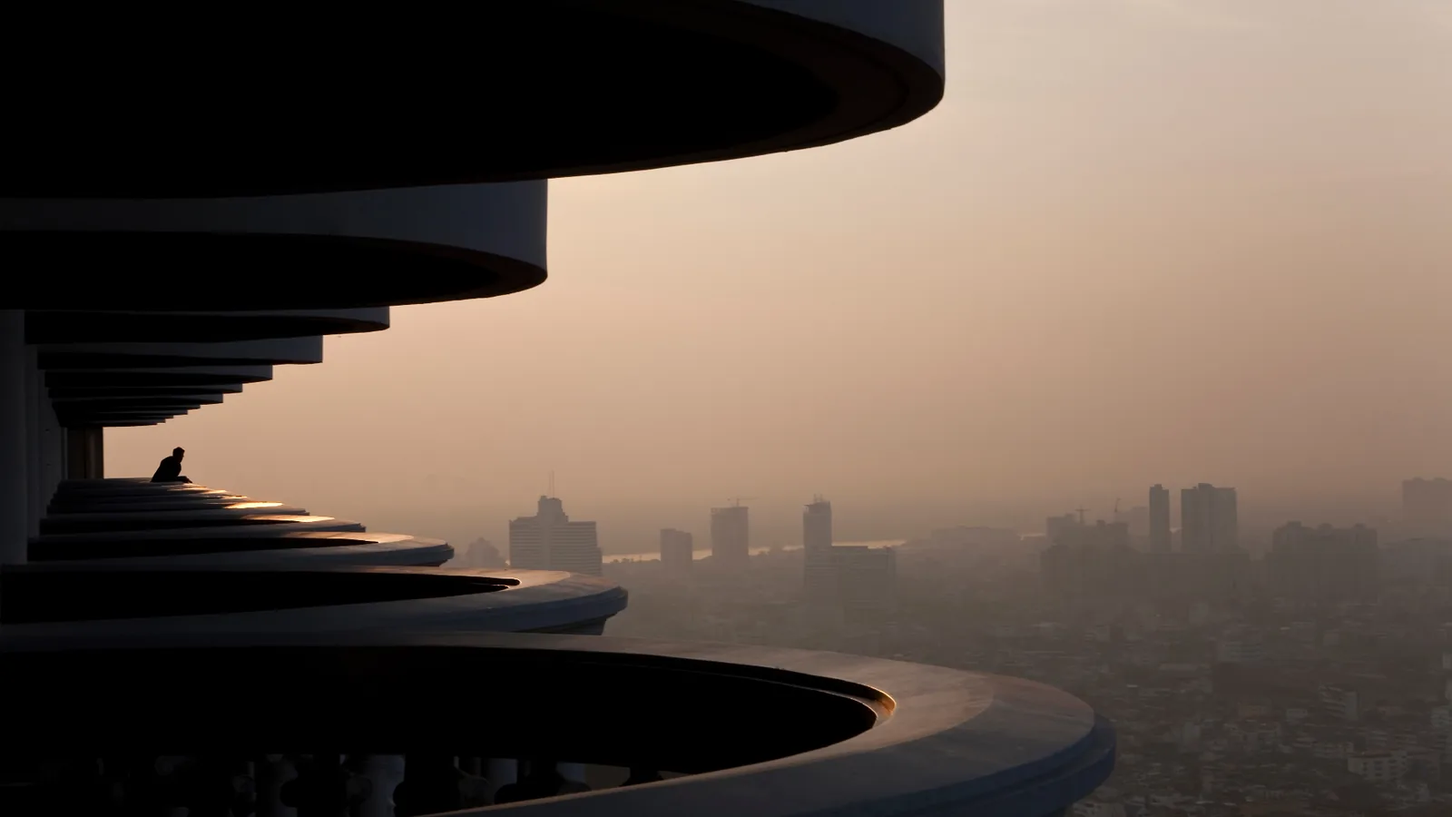 Curved skyscraper balconies overlooking a hazy city skyline at dusk, with a lone figure seated on the ledge